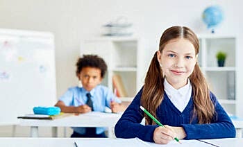 Drawing by desk Adorable schoolgirl with crayon sitting by desk with classmate on background